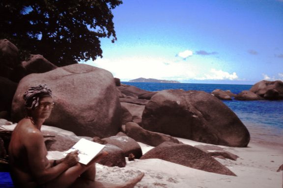 Beim Malen an der Anse Lazio Person sitzt am Strand und liest, umgeben von großen Felsen und dem Meer im Hintergrund.