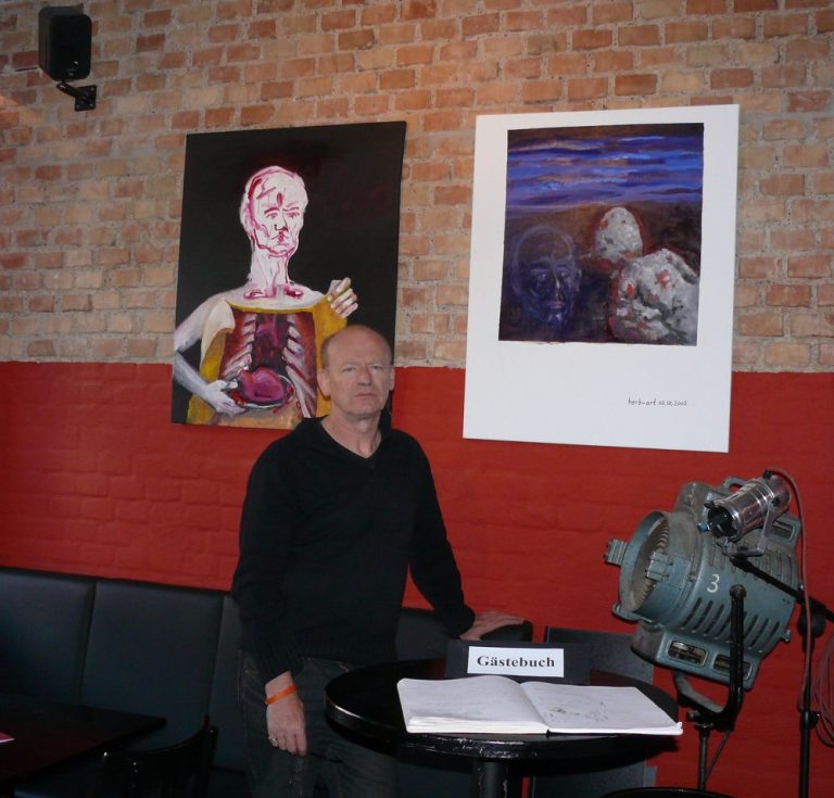 Mann steht neben Kunstwerken in einem modernen Raum mit Backsteinwand und rotem Bereich.